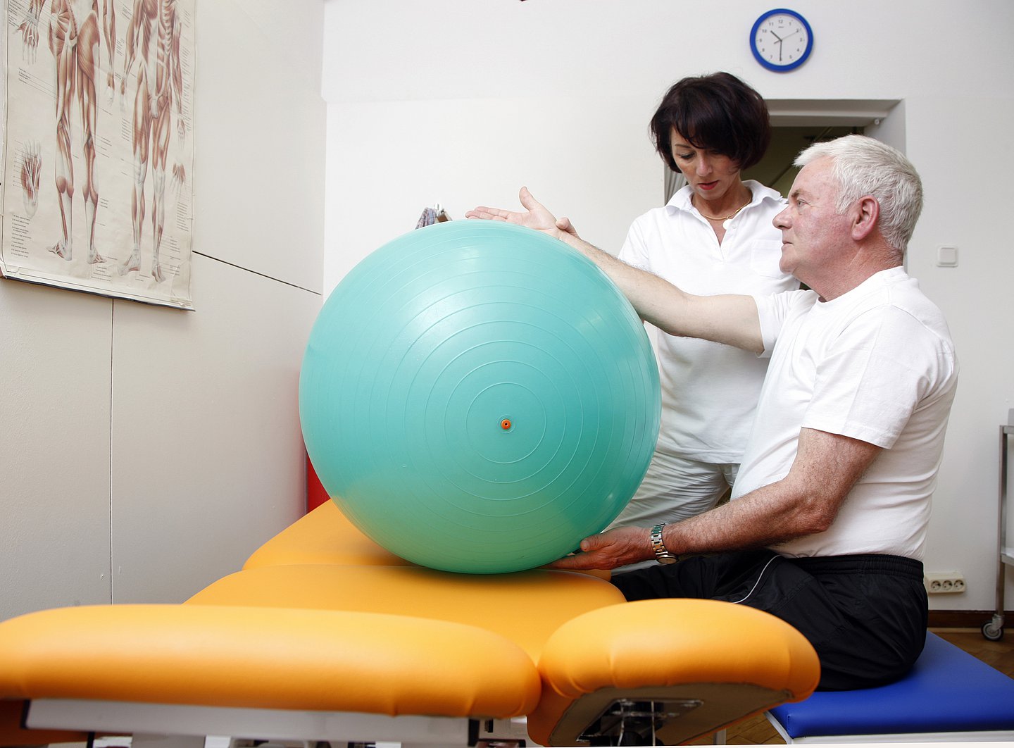 Eine Physiotherapeutin (stehend) unterstützt einen Senior (sitzend) bei der Physiotherapie an einem großen Gymnastikball
