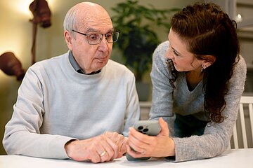 Ein &auml;lterer Mann sitzt an einem Tisch, w&auml;hrend eine l&auml;chelnde Frau ihm ein Smartphone zeigt und ihm dessen Funktionen erkl&auml;rt.