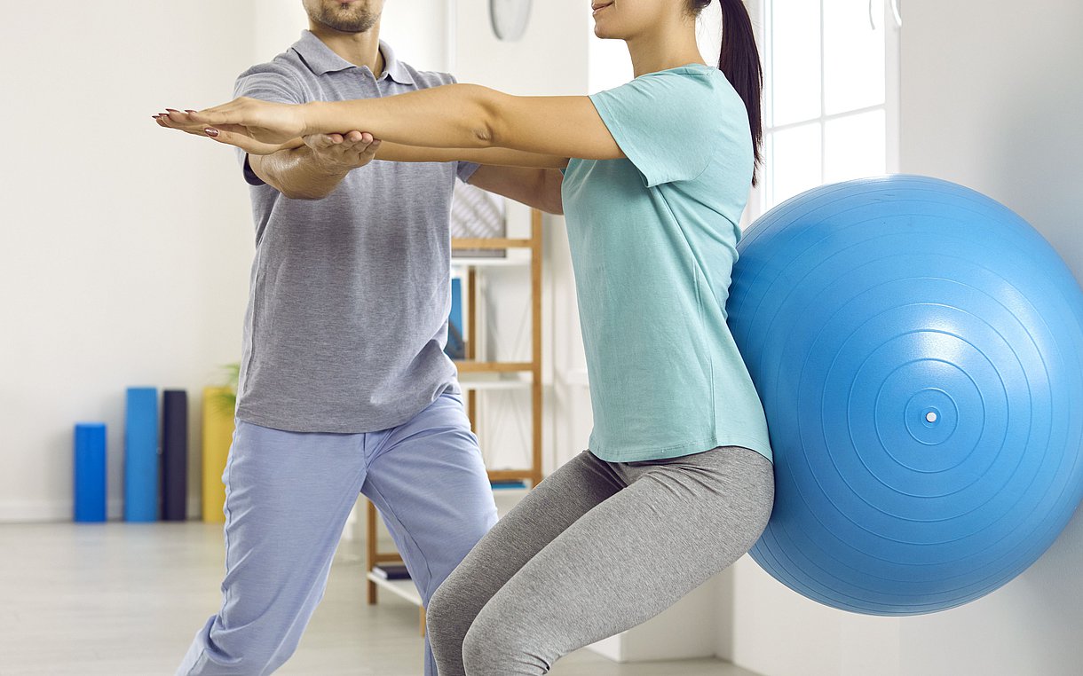 Foto: Ein Physiotherapeut korrigiert die Rückenübung einer Patientin mit einem Gymnastikball.