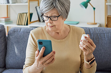 Eine Frau in Nahaufnahme sitzt auf der Couch mit einer Tablettendose in der Hand und schaut auf ihr Handy.