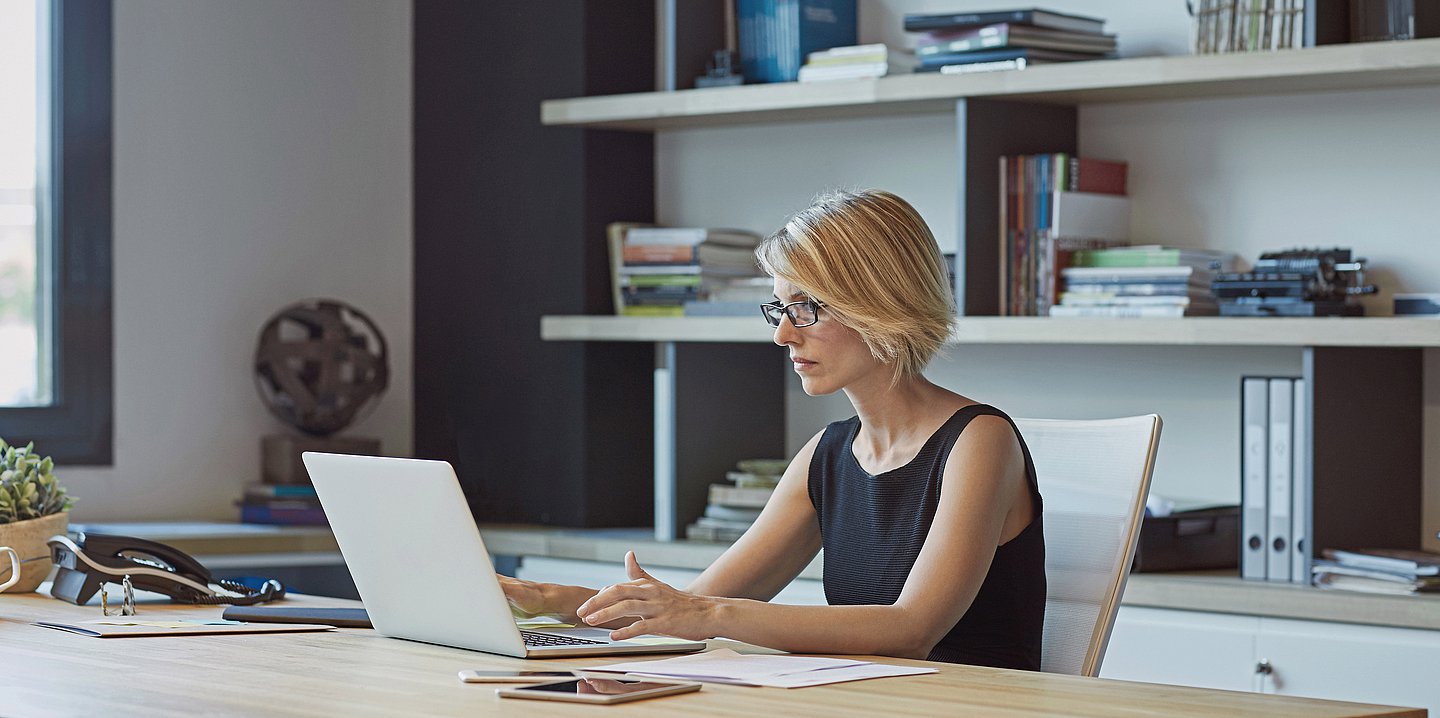 Eine Frau sitzt am Schreibtisch und arbeitet an einem Laptop.