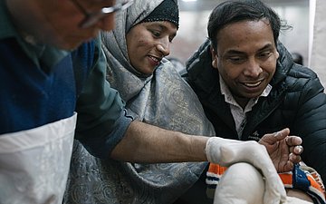 Ein Baby bekommt seinen ersten Gips gegen Klumpfüße. Die Eltern stehen neben dem Arzt, der den Gips anlegt und schauen das Kind aufmunternd an.