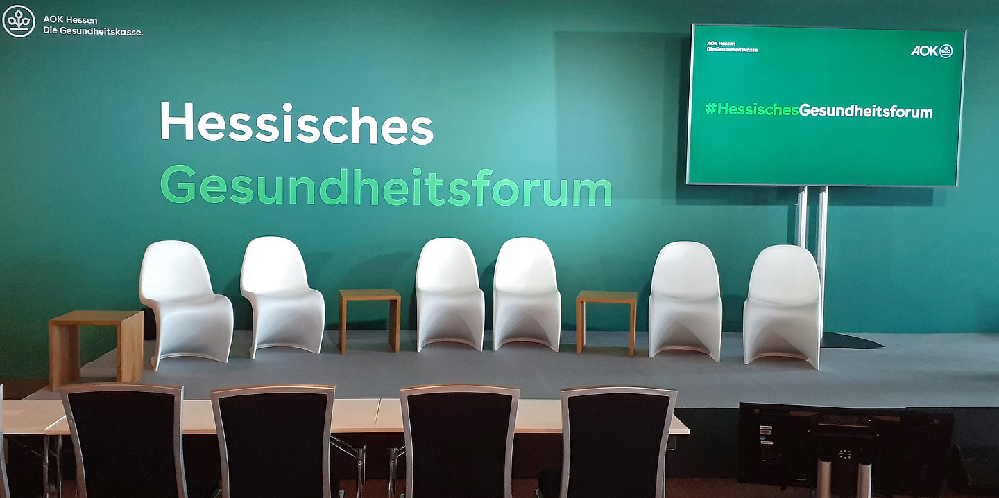 Das Foto zeigt ein Podium mit sechs weißen Stühlen vor einer grünen Wand mit der Aufschrift Hessisches Gesundheitsform. 