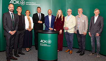 Eine Gruppe steht vor einem gr&uuml;nem Hintergrund. Davor steht ein runder Tisch mit Logo an der Vorderseite.