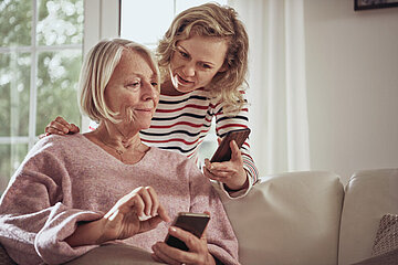 Eine &auml;ltere Frau sitzt auf einem Sofa. Schr&auml;g hinter ihr beugt sich eine j&uuml;ngere Frau, vermutlich ihre Tochter, &uuml;ber sie. Beide gucken auf ihr Smartphone. Die Szene wirkt, als w&uuml;rde die j&uuml;ngere der &auml;lteren Frau etwas erkl&auml;ren.