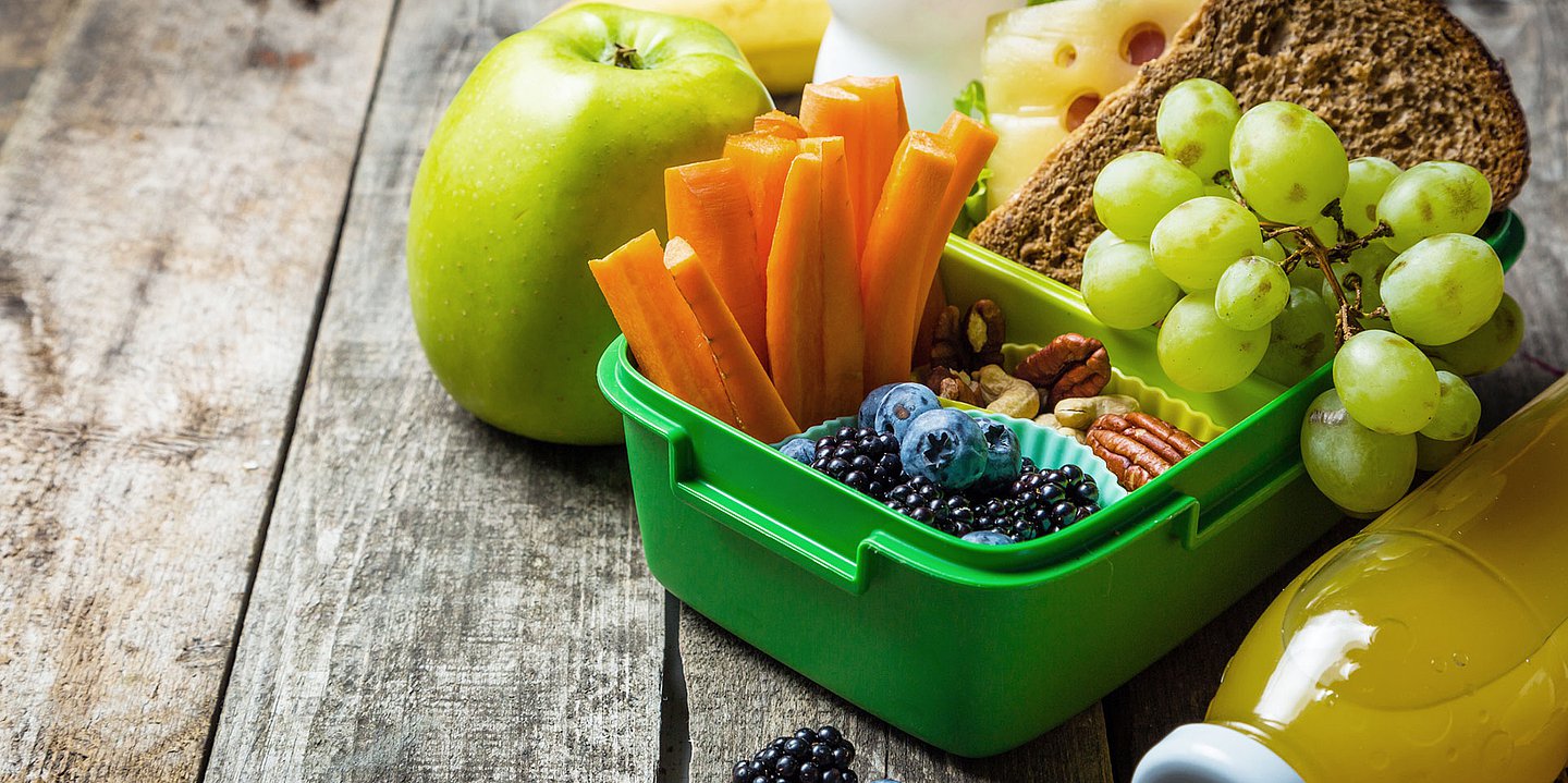 Foto: Eine Früstücksbox mit Obst, Gemüse und Vollkornbrot steht geöffnet auf einem Tisch, daneben liegen ein Apfel und eine Flasche Orangensaft.