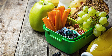 Foto: Eine Fr&uuml;st&uuml;cksbox mit Obst, Gem&uuml;se und Vollkornbrot steht ge&ouml;ffnet auf einem Tisch, daneben liegen ein Apfel und eine Flasche Orangensaft.