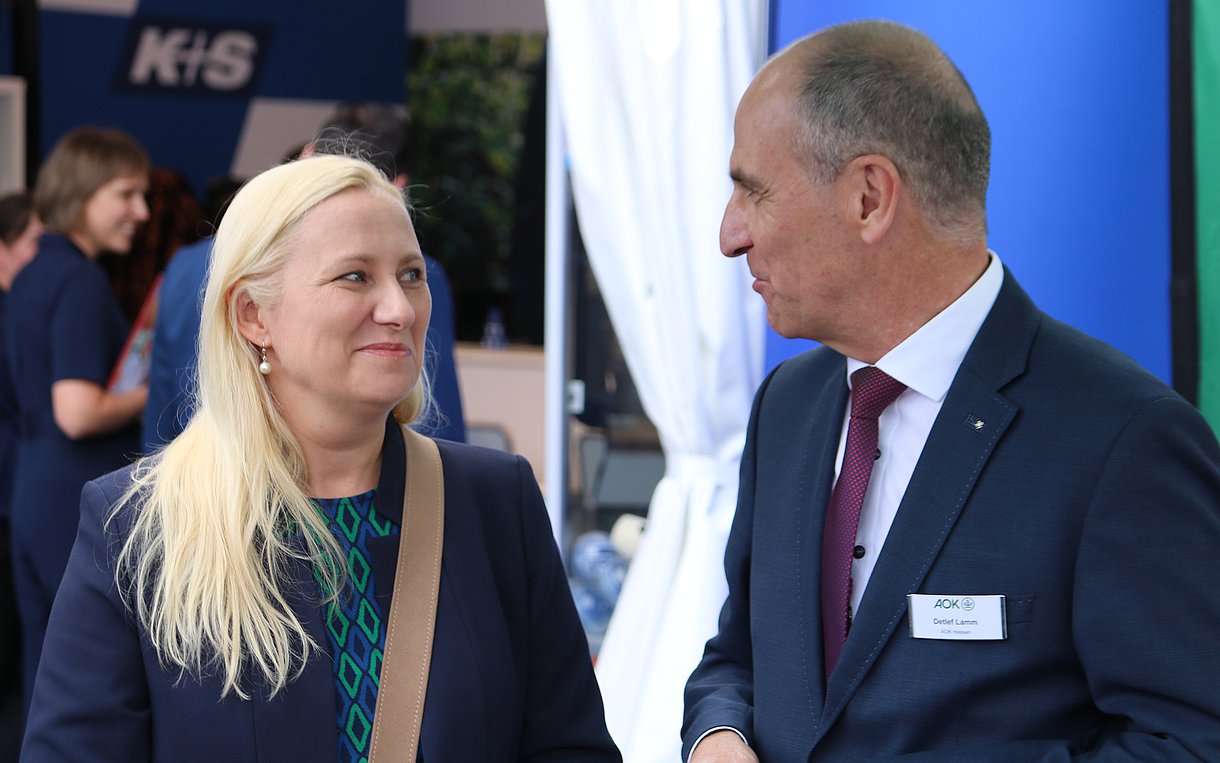 Das Foto zeigt auf der linken Seite die hessische Gesundheitsministerin Diana Stolz und rechts daneben den Vorstandsvorsitzenden der AOK Hessen, Detlef Lamm, im Gespräch. Diana Stolz hat lange hellblonde Haare und trägt einen blauen Blazer, darunter eine grün gemusterte Bluse. Detlef Lamm trägt einen dunkelblauen Anzug mit weißem Hemd und rot-blau gestreifter Krawatte.