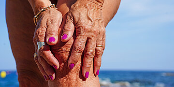 Foto einer Frau am Strand, die sich das Knie h&auml;lt. Es sind nur Teile ihrer Beine und ihre H&auml;nde mit Nagellack zu sehen.