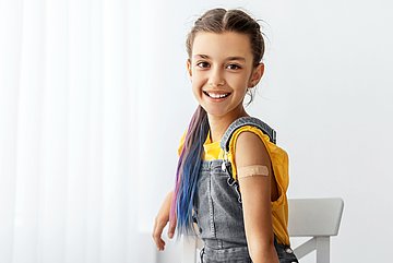 Ein junges Mädchen sitzt nach der HPV-Impfung auf einem Stuhl, hat ein Pflaster am Arm und lächelt