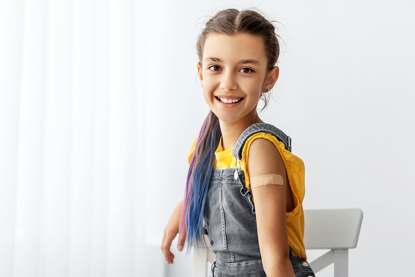 Ein junges Mädchen sitzt nach der HPV-Impfung auf einem Stuhl, hat ein Pflaster am Arm und lächelt