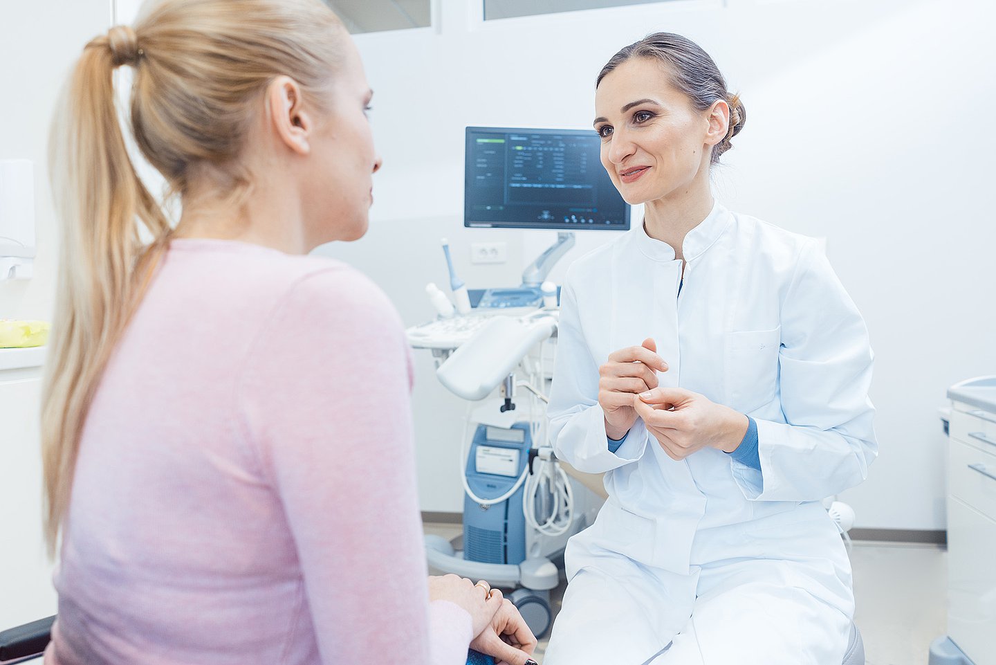 Eine Ärztin sitzt auf einem Hocker in ihrem Behandlungszimmer und hat sich einer Patienten zugewandt, mit der sie im Gespräch ist.