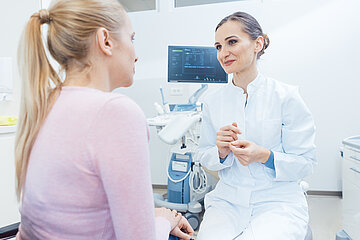 Eine &Auml;rztin sitzt auf einem Hocker in ihrem Behandlungszimmer und hat sich einer Patienten zugewandt, mit der sie im Gespr&auml;ch ist.