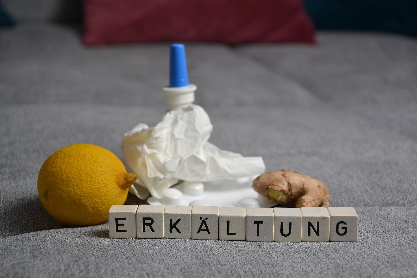 Kleine Holzwürfel mit den Buchstaben 'Erkältung', eine Zitrone, ein Papiertaschentuch, eine Flasche Nasenspray und ein Stück Ingwer liegen in Nahaufnahme auf einem Teppich.