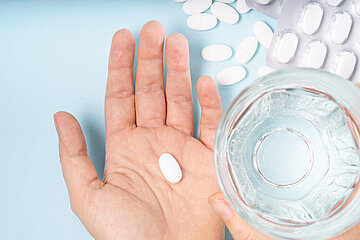 Auf einer Handinnenfl&auml;che liegt eine Tablette, daneben steht ein Glas Wasser, daneben liegen weitere Tabletten.