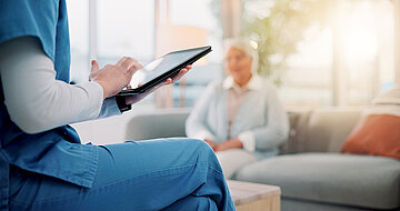 Im Vordergrund sitzt eine Pflegekraft mit einem Tablet in der Hand, im Hintergrund und nur verschwommen sichtbar, sitzt eine Ältere Person, die befragt wird.
