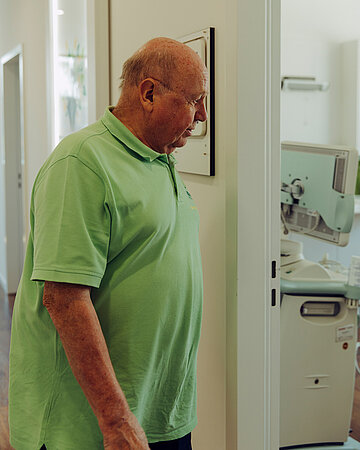 Foto: Peter Engeser steht in einer Arztpraxis vor einem Untersuchungsraum und blickt auf ein medizinisches Ger&auml;t.