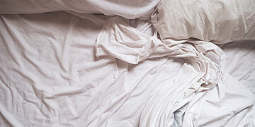 Draufsicht auf ein ungemachtes Bett mit wei&szlig;en, zerknitterten Bettlaken und Kissen. Die Bettw&auml;sche liegt locker in weichen Falten und ist in sanftes, nat&uuml;rliches Licht getaucht.