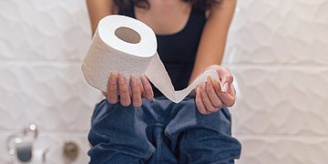Toilettengang Foto: Eine junge Frau sitzt und hat Toilettenpapier in der Hand.