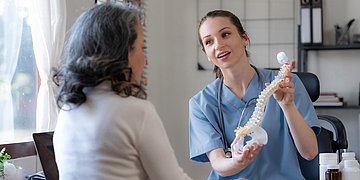 Eine ältere Frau sitzt im Besprechungszimmer. Vor ihr sitzt eine junge Ärztin und hält ein Modell eine Wirbelsäule hoch.