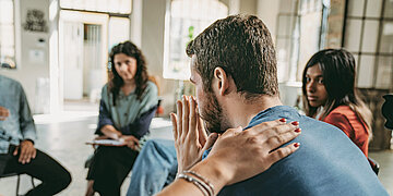 Foto: Einige Menschen sitzen in einem Stuhlkreis, ein Mann h&auml;lt sich die H&auml;nde vor das Gesicht, w&auml;hrend eine andere Hand tr&ouml;stend auf seinem R&uuml;cken liegt.