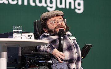 Ra&uacute;l Krauthausen sitzt in einem Rollstuhl, der auf der B&uuml;hne steht. Er spricht in ein Mikrofon. 