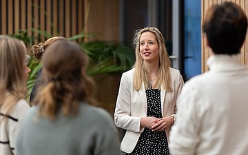 Eine Beratungs-Szene mit einer jungen Frau im weißen Sacko, die mit drei Personen spricht. Alle vier stehen in einem Raum mit Holzvertäfelung und größeren Grünpflanzen.