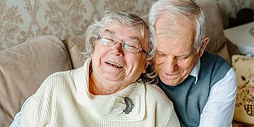 Zwei ältere Menschen sitzen auf einem Sofa. Der Mann umarmt von hinten die Frau. Beide lächeln.