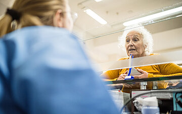&Auml;ltere Frau mit wei&szlig;en Haaren steht an einem Serviceschalter hinter einer Glasscheibe und spricht mit einer Person in blauer Kleidung, die im Vordergrund von hinten zu sehen ist.