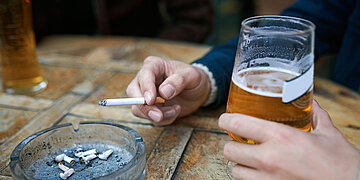 Foto einer Person, die an einem Holztisch sitzt mit Bierglas in der Hand und Zigarette in der anderen. Vor ihr steht ein Aschenbecher. Es ist nur der Berieich der H&auml;nde zu sehen.
