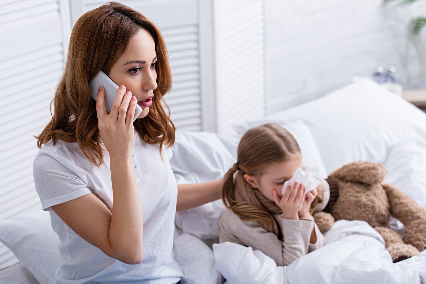 Foto zeigt eine Mutter, die am Bett ihres kranken Kindes sitzt und ein Telefongespräch führt.