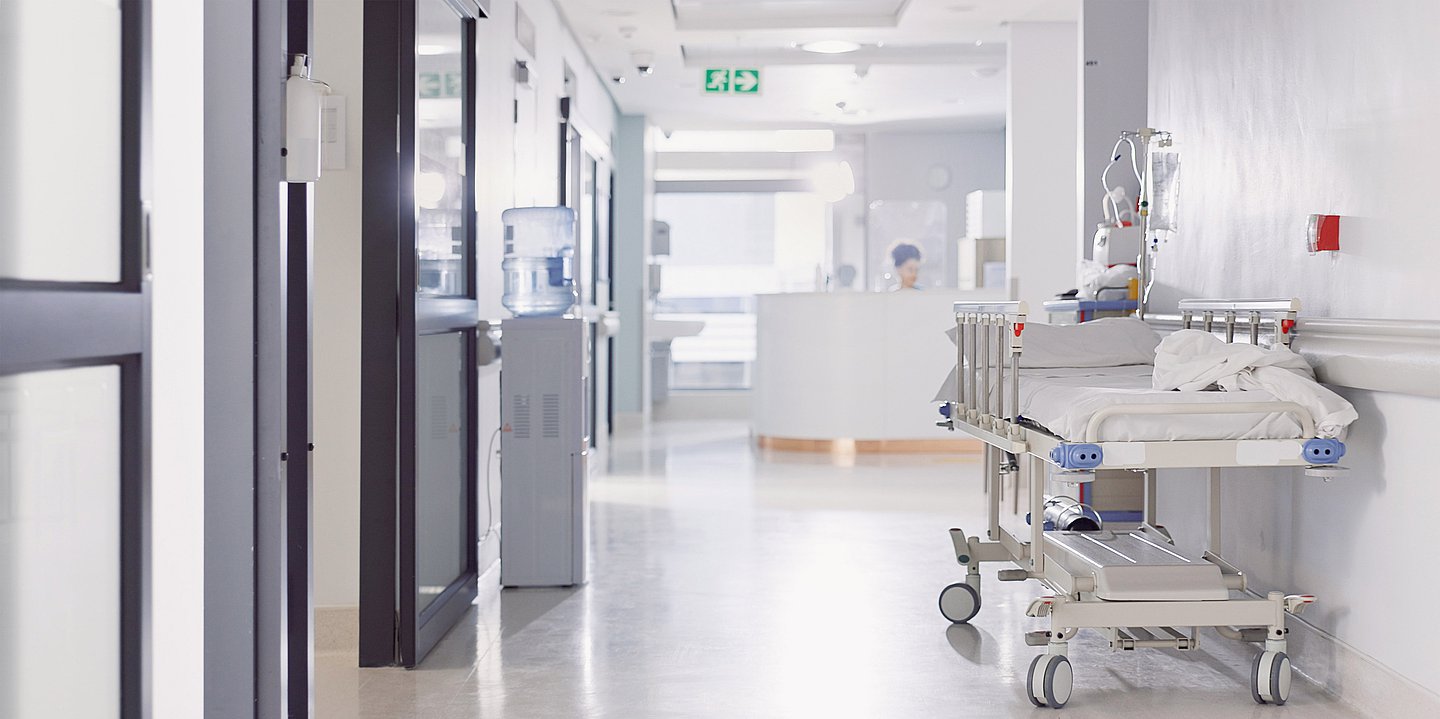 Ein leerer Krankenhausflur, am rechten Rand steht ein leeres Krankenhausbett. Ganz hinten ein Tresen, hinter dem eine Frau sitzt und arbeitet.