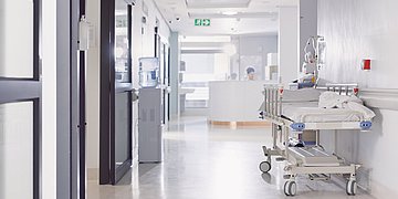 Krankenhausreform Ein leerer Krankenhausflur, am rechten Rand steht ein leeres Krankenhausbett. Ganz hinten ein Tresen, hinter dem eine Frau sitzt und arbeitet.