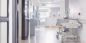Ein leerer Krankenhausflur, am rechten Rand steht ein leeres Krankenhausbett. Ganz hinten ein Tresen, hinter dem eine Frau sitzt und arbeitet.