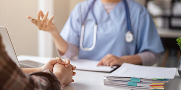 Arzt und Patient sind im Gespr&auml;ch. Beide sind nur im Anschnitt abgebildet. Sie sitzen auf zwei Seiten eines Tisches, einander zugewandt. In der Mitte des Tisches liegt eine Patientenakte.