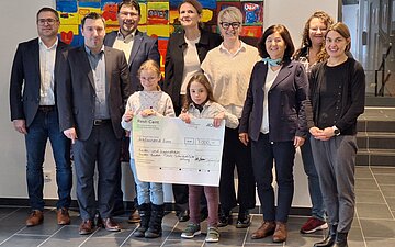 Gruppenfoto zur Scheck&uuml;bergabe der Rest-Cent-Initiative 2026: In der Mitte halten zwei Kinder einen gro&szlig;formatigen Scheck.