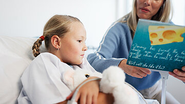 Ein M&auml;dchen liegt im Krankenhausbett, am Finger ein Messger&auml;t und im Arm ein Schmusetier. Am Bett sitzt eine Frau und liest ihr aus einem Buch vor.