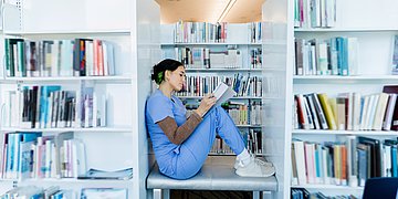 Eine Frau in medizinischer Berufskleidung sitzt zwischen großen Bücherregalen und liest ein Buch.