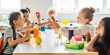 Zwei junge M&auml;dchen l&auml;cheln und teilen sich ein Sandwich an einem Schultisch, im Hintergrund sitzen weitere Kinder mit bunten Getr&auml;nken.