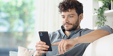 Auf einer wei&szlig;en Couch sitzt ein Mann. Ein Arm liegt auf der Lehne. Er schaut in sein Smartphone.