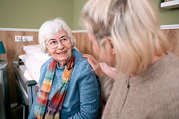 Eine Seniorin sitzt auf der Bettkante ihres station&auml;ren Bettes. Vor ihr, im unscharfen Vordergrund, sitzt eine j&uuml;ngere Frau und hat die Hand auf der Schulter der Seniorin.