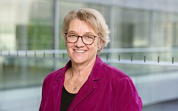 Das Bild zeigt eine l&auml;chende blonde Brillentr&auml;gerin (Oberk&ouml;rper) mit k&uuml;rzeren gewellten Haaren. Sie tr&auml;gt einen weinroten Jeans-Blazer und ein schwarzes Top. Sie tr&auml;gt ein Halskettchen mit rundem Anh&auml;nger und steht in einem gl&auml;sernen B&uuml;rogeb&auml;ude (unscharfer Hintergrund)