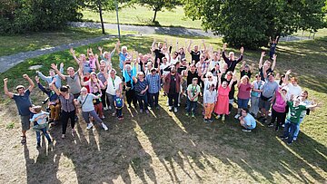 Das Foto zeigt eine bunte Menschen Gruppe auf einer Wiese in einer Parkanlage schr&auml;g von oben. Die Sonne scheint und die Gruppe wirft einen gro&szlig;en Schatten nach vorne. Im Hintergund sind B&auml;ume zu sehen.