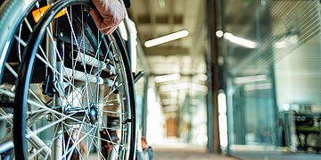 Foto: Rad eines Rollstuhls, eine Hand greift das Rad, dahinter ein langer Flur.
