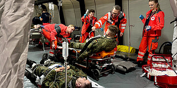 Sanit&auml;ter in roten Uniformen versorgen verletzte Soldaten auf Tragen in einem Feldlazarett, umgeben von medizinischer Ausr&uuml;stung.