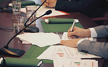 Konferenz Foto: Mikro und viele Unterlagen auf einem Tisch, dazu schreibt jemand etwas auf Blätter mit Diagrammen