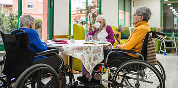 An einem runden Tisch sitzen drei &auml;ltere Personen im Rollstuhl. Eine Frau trinkt aus einer Tasse, ein Mann schreibt etwas auf. Die dritte blickt aus dem Fenster.