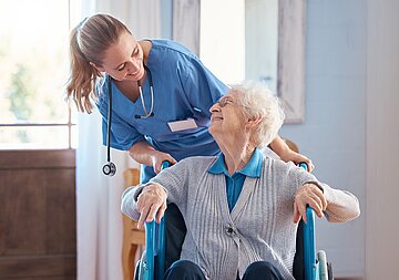 Eine Pflegekraft schiebt eine Pflegebed&uuml;rftige im Rollstuhl. Dabei unterhalten sich die beiden.