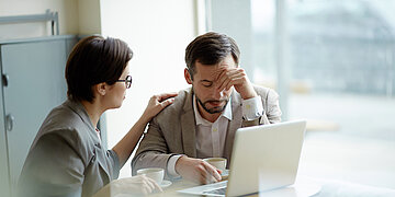 Ein Mann sitzt mit verzweifeltem Gesichtsausdruck vor einem Laptop, eine Frau legt ihm f&uuml;rsorglich die Hand auf die Schulter.