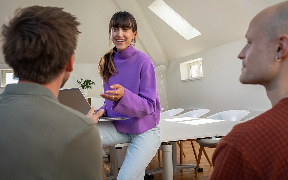 eine junge Frau sitzt auf einem Tisch und unterhält sich mit zwei Kollegen.